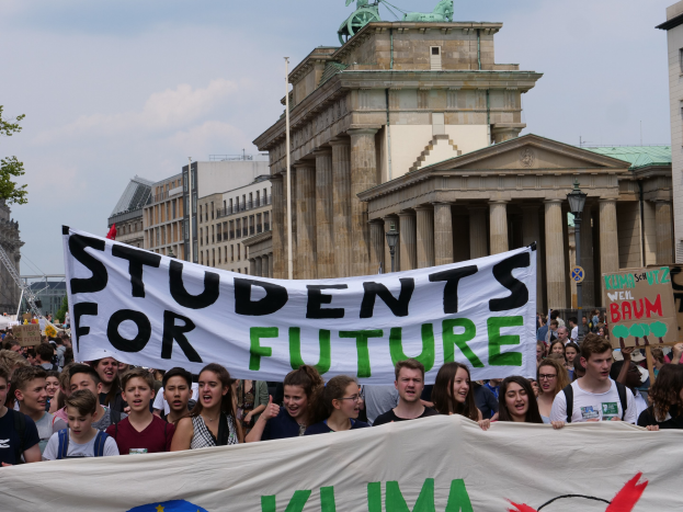 Eine Gruppe von Schülern marschiert in Berlin, die eine bunt bemalte "Students for Future"-Tafel trägt, vor einer Kulisse aus Gebäuden, Bäumen und Himmel.