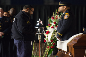 Zwei Polizeibeamte in Uniform stehen feierlich neben einem mit Blumen geschmückten Sarg, mit einigen Menschen im Hintergrund.