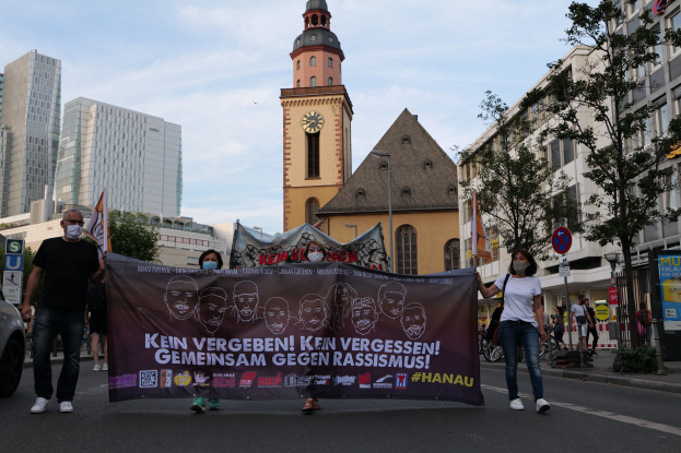 Eine Gruppe von Menschen in Masken, die eine Straße entlangmarschieren und ein Banner halten, mit einem geparkten Auto auf der linken Seite, Gebäuden und Bäumen im Hintergrund und einem Uhrenturm unter einem klaren blauen Himmel.