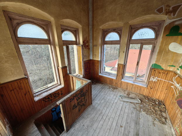 Ein verlassenes Gebäude mit Graffiti an den Wänden und Fenstern, einem Holzboden, einer Treppe und verstreuten Gegenständen, mit Bäumen und einem Haus im Hintergrund.