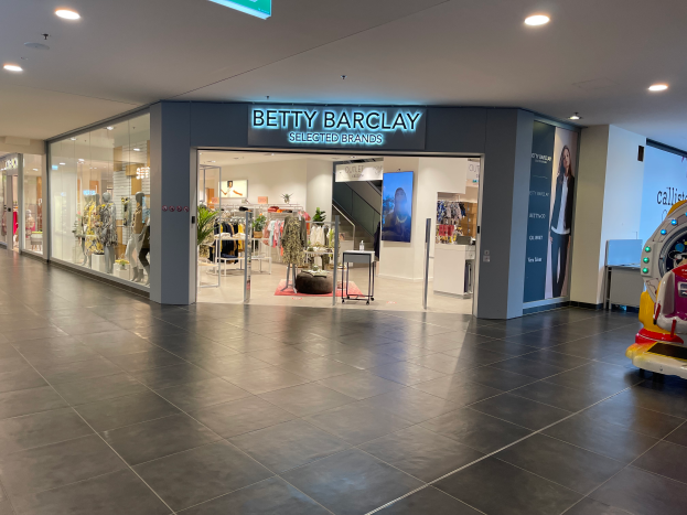 Das Innere eines Betty Barclay-Geschäfts in einem Einkaufszentrum, das Schaufensterpuppen in Kleidern, hängende Kleidung, Tische, Pflanzen und andere Gegenstände zeigt, mit einem Spielzeugfahrzeug auf der rechten Seite und einer Tafel mit der Aufschrift "Betty Barclay Selected Brands" oben.