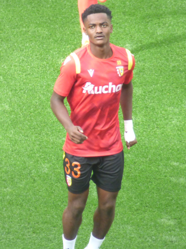 Emmanuel Adebayor in rotem T-Shirt und schwarzen Shorts auf einem Fußballfeld mit zwei Personen im Hintergrund.