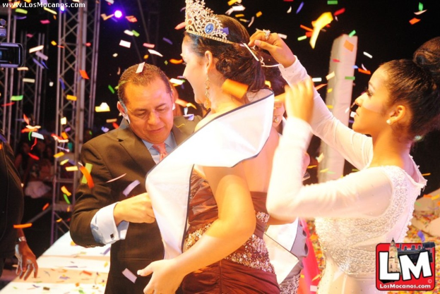 Ein Mädchen auf der Bühne bei einer Miss World Show mit einer Krone, mit einem anderen Mädchen, das sie hinter ihr anpasst, und einem Mann in einem schwarzen Anzug, der vor ihr die Hand schüttelt, vor einem dunklen Hintergrund mit Metallrahmen.