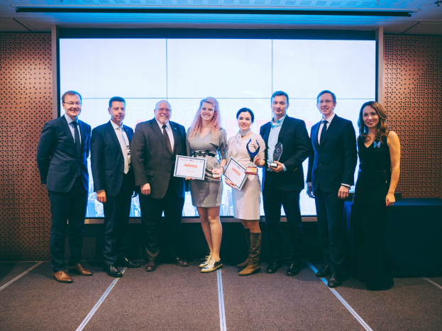 Gruppe von strahlenden Menschen mit Urkunden vor einem Bildschirm, mit einem Tisch und einem Wandobjekt im Hintergrund, Gewinner der Internationalen Business Awards 2018.