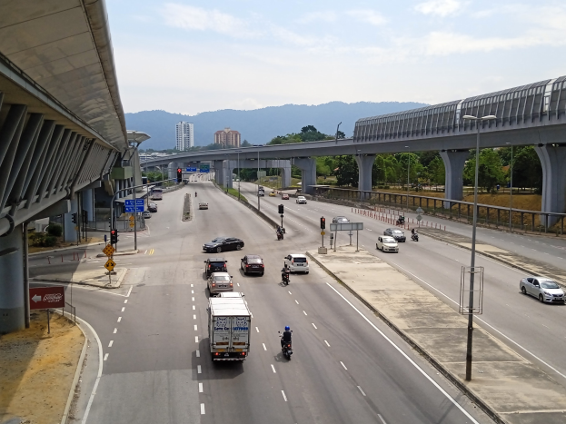 Eine belebte Autobahn mit mehreren Fahrzeugen in der Nähe einer Brücke, mit Straßenlaternen, Verkehrszeichen, Schildern, Bäumen, Gebäuden, Hügeln und einem bewölkten Himmel.