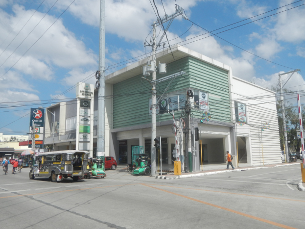 Eine belebte Straße mit Fahrzeugen, Fußgängern, Strommasten, Gebäuden, Bäumen und einem bewölkten Himmel, mit einer Tankstelle mit parkenden Autos im Vordergrund.