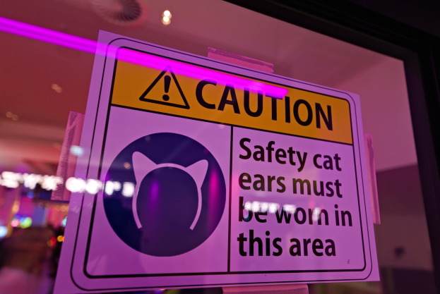 Hellgelbes Warnschild in einem Ladenschaufenster mit schwarzer Schrift, die "Achtung: Sicherheitskatzenohren müssen in diesem Bereich getragen werden" lautet.