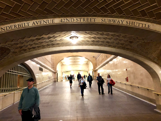 Eine Gruppe von Menschen geht durch eine belebte U-Bahn-Station in New York City, mit einem Gel├Ąnder auf der linken Seite, Lampen an der Decke und Text an den W├Ąnden.
