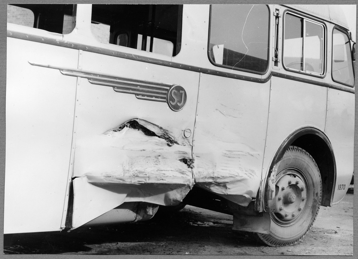 Schwarzes und weißes Foto eines auf der Straße gestrandeten Busses mit eingedellter Front, zersplitterten Fenstern und geöffneter Fahrertür, die den Innenraum zeigt.