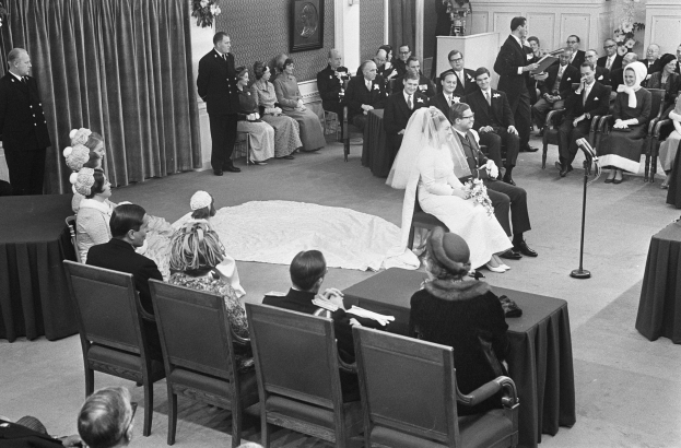 Schwarzes und weißes Foto einer Hochzeitszeremonie mit sitzenden und stehenden Menschen, einem Mann mit Mikrofon und einer Frau mit einem Bukett, mit einem Rednerpult, Blumenschmuck, einem Vorhang, einem gerahmten Foto an der Wand und einer Tür im Hintergrund.