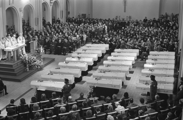 Eine große Gruppe von Menschen, die in einer Kirche sitzen, mit einigen, die davor stehen, ein Podium mit einem Mikrofon links daneben und Blumensträuße mit Lichtern im Hintergrund, alles in Schwarz-Weiß.