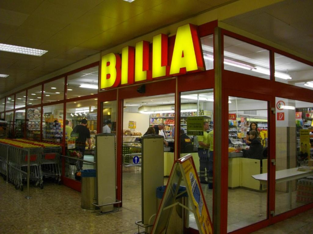 Ein Markt mit einem Eingang in der Mitte, Mülltonnen daneben, einer Reihe von Einkaufswagen auf der linken Seite, Deckenbeleuchtung und einigen Menschen, die im Inneren herumlaufen.