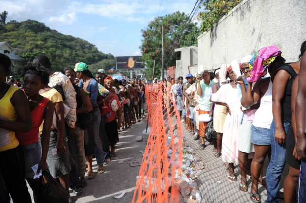 Lange Schlange von Menschen, die an einer Wahllokal-Schranke warten, mit einem Zaun, einer Wand, Bäumen, Gebäuden, Strommasten und einer Werbetafel im Hintergrund.