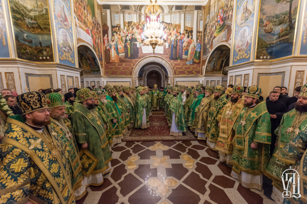 Eine Gruppe von Priestern in grünen und goldenen Gewändern und Mützen steht in einer Kirche mit Gemälden und einem Kronleuchter, mit einem Bogen und einigen Menschen im Hintergrund, die den St. Peters Tag feiern.