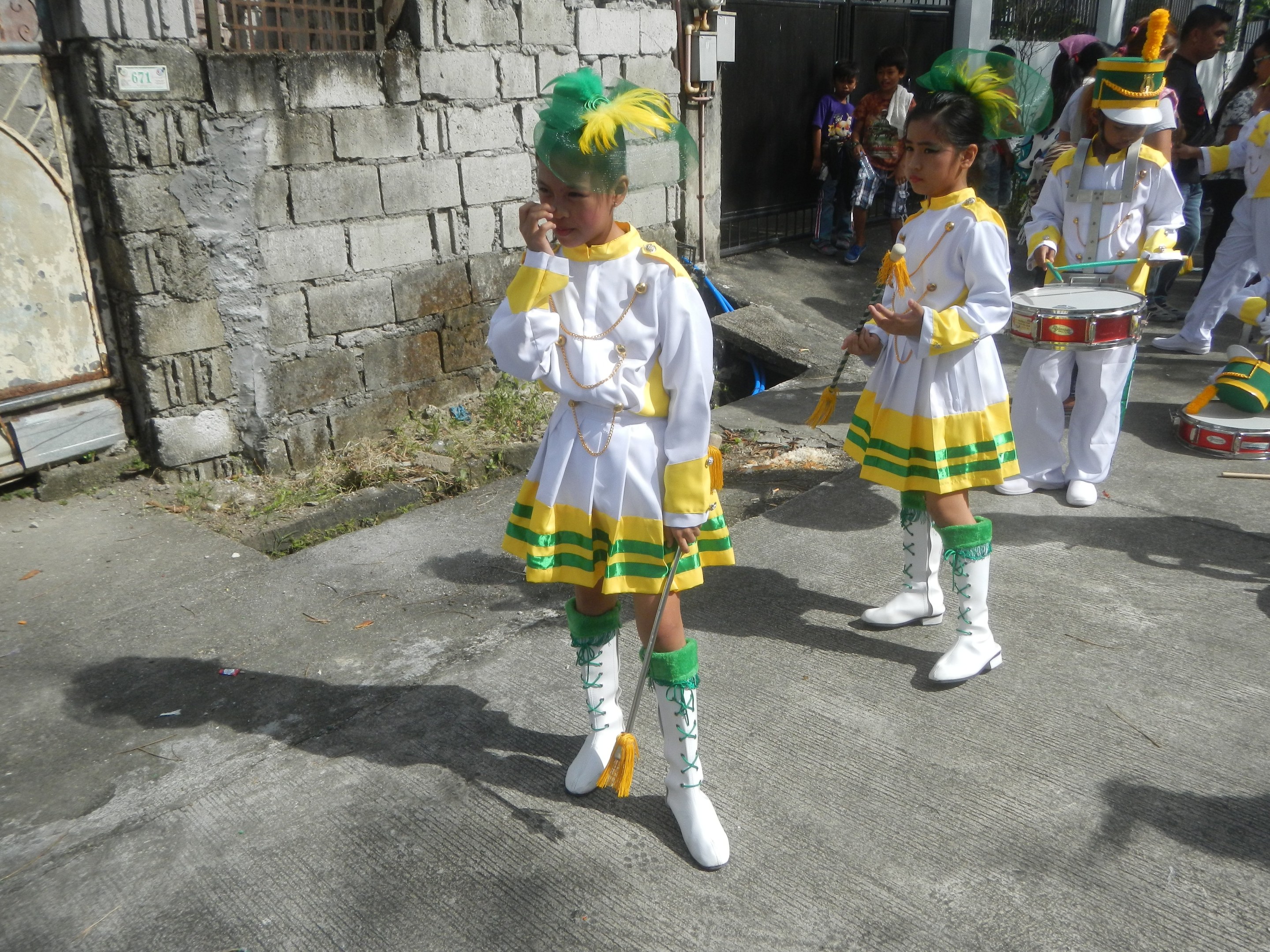 Gruppe junger Mädchen in weißen und gelben Outfits, die marschieren und musikalische Instrumente spielen, mit einer Wand, einem Tor und Bäumen im Hintergrund.