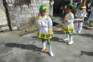 Gruppe junger Mädchen in weißen und gelben Outfits, die marschieren und musikalische Instrumente spielen, mit einer Wand, einem Tor und Bäumen im Hintergrund.