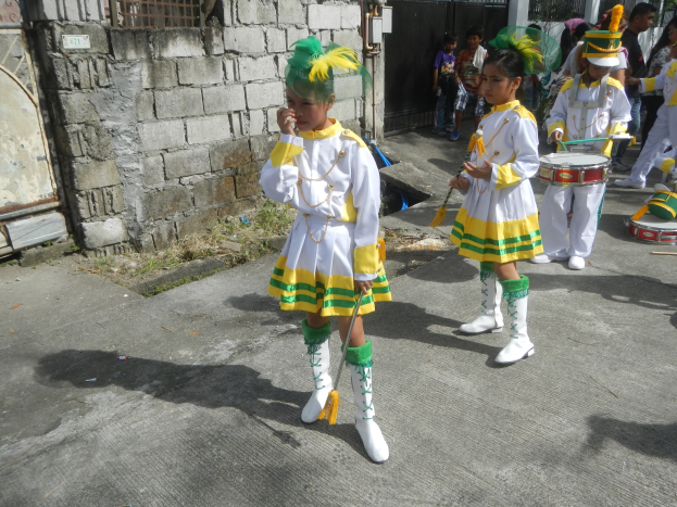 Gruppe junger Mädchen in weißen und gelben Outfits, die marschieren und musikalische Instrumente spielen, mit einer Wand, einem Tor und Bäumen im Hintergrund.