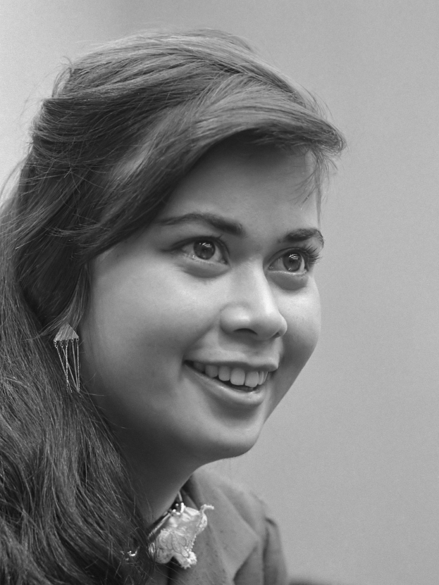 Ein Schwarz-Weiß-Foto einer Frau mit langen Haaren, die zu einem ordentlichen Knoten frisiert sind, die warm in die Kamera lächelt und direkt in die Augen des Betrachters schaut.