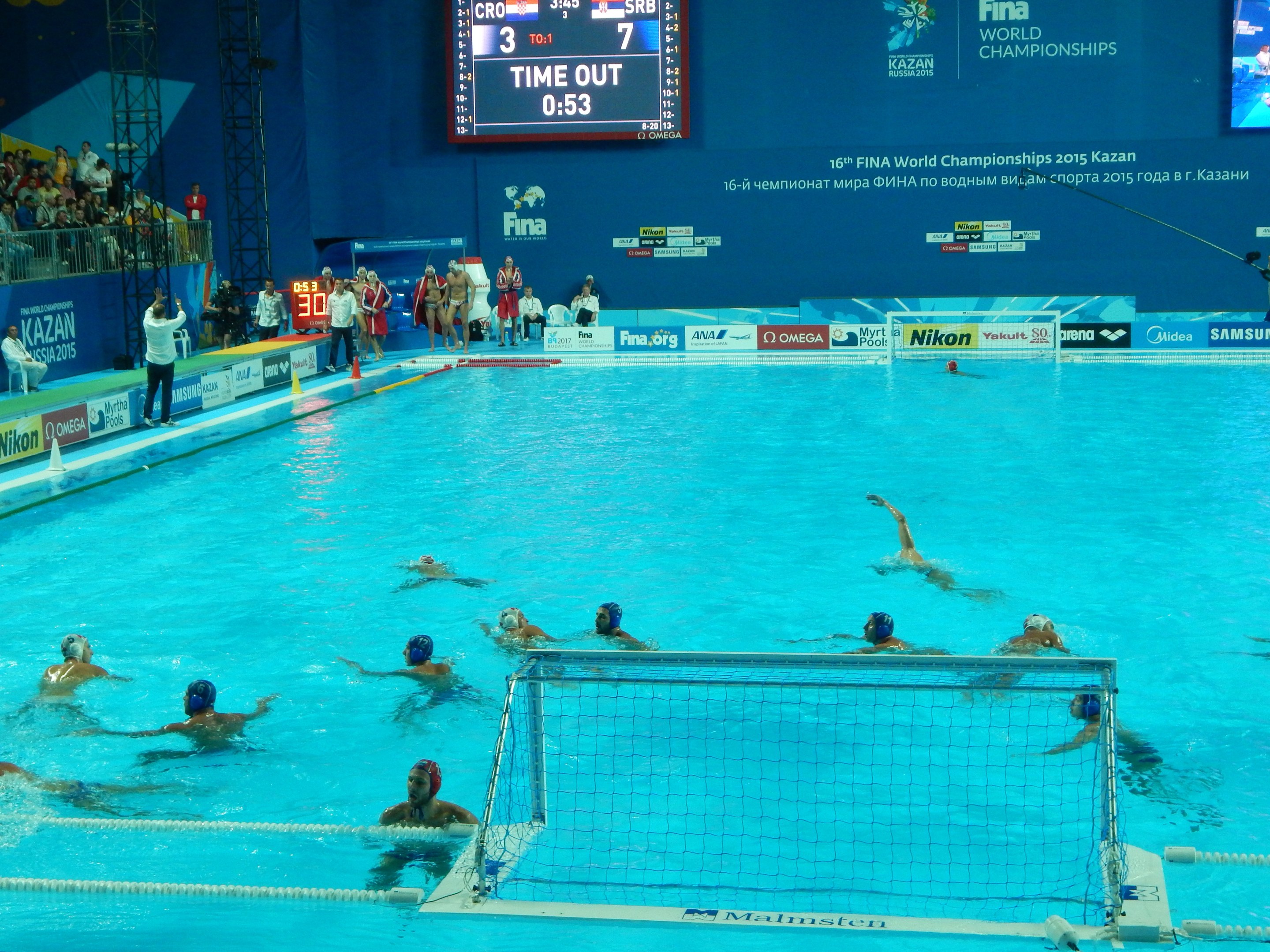 Water polo players competing in a pool during the 2015 FINA World Championships in Kazan, with spectators and scoreboards visible in the background.
