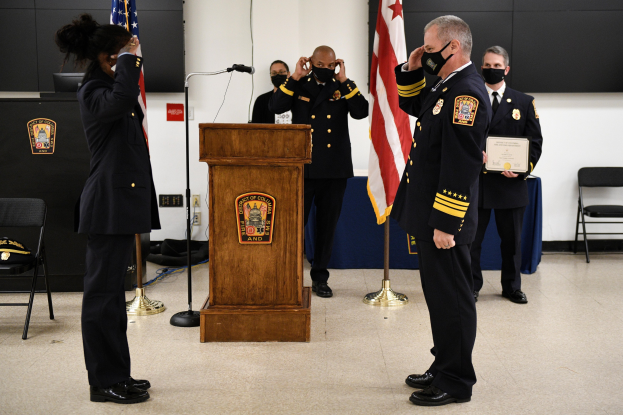 Gruppe uniformierter Personen in Masken vor einem Rednerpult bei einer Vereidigung eines neuen Feuerwehrchefs, der eine Urkunde hält, mit Fahnen, Stühlen, Tischen und einer Wand im Hintergrund.