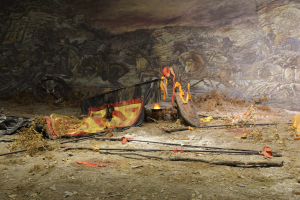 Ein Gemälde an einer Gebäudewand mit einem brennenden Feuer davor, umgeben von verstreuten Kleidungsstücken, Stöcken und anderen Gegenständen, mit einem Rad im Vordergrund.