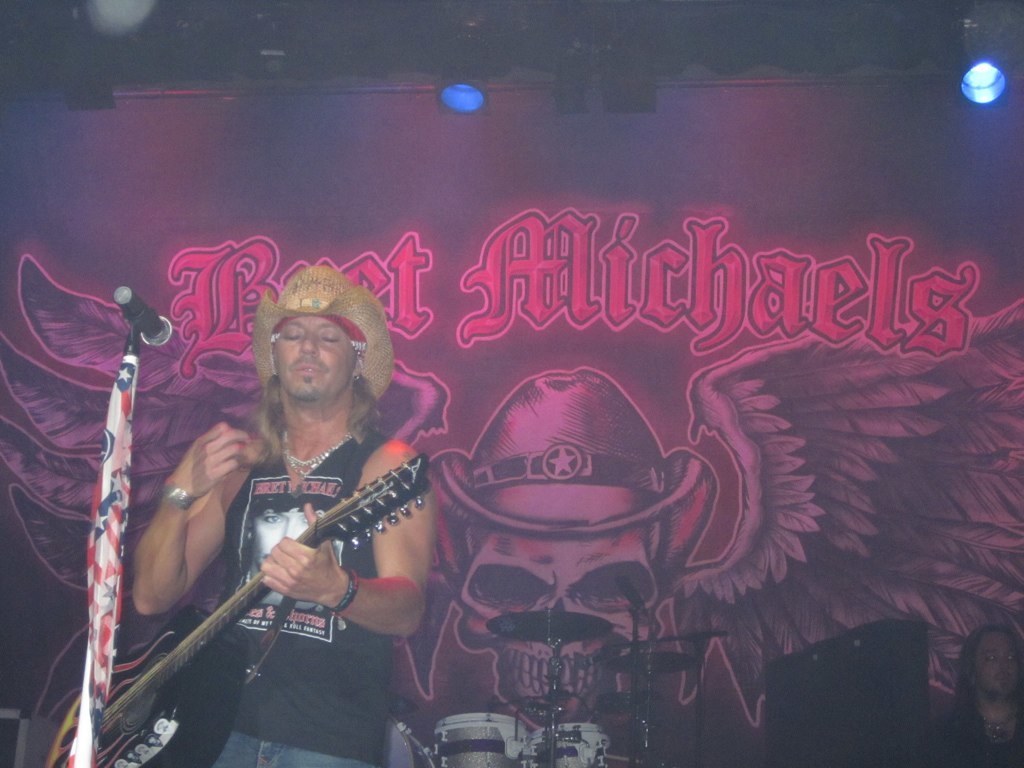 Eine Musikkonzertszene mit Scheinwerferlicht oben, einem Banner im Hintergrund, einer sitzenden Person in der rechten unteren Ecke, einem stehenden Gitarristen links mit einem Mikrofon davor und Gegenständen in der Mitte unten.