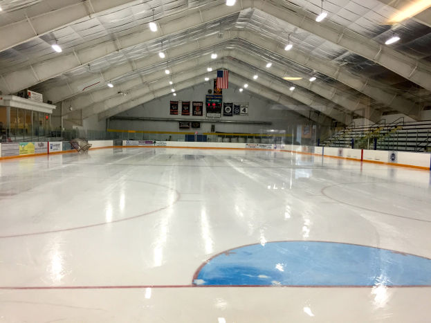 Eine große Indoor-Eisfläche mit Deckenbeleuchtung, bedruckten Tafeln, einer Hintergrundwand, Treppen mit Geländern und einigen Menschen, wahrscheinlich eine Hockey-Einrichtung.