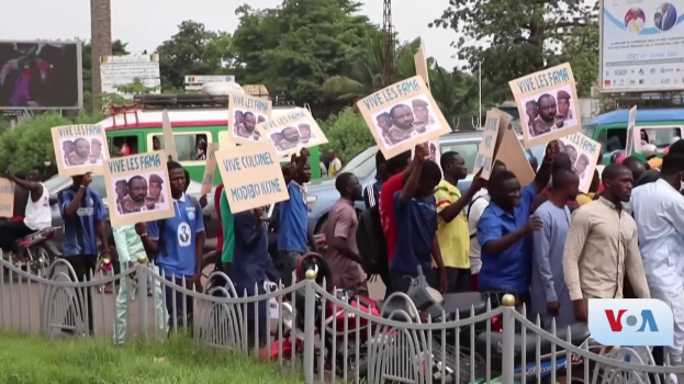 Eine Gruppe von Menschen mit Schildern steht vor einem Zaun, umgeben von Fahrzeugen, Bäumen, Masten, Werbetafeln und einem Bildschirm, mit Gras unten und dem Himmel im Hintergrund, scheinbar protestieren sie gegen eine Regierungentscheidung, die Video-Konferenzen verbietet.