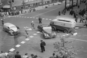 Schwarzes und weißes Bild eines Autounfalls an einer Straßenkreuzung mit mehreren Fahrzeugen, darunter ein Lieferwagen, und einer Gruppe von Schaulustigen; Gebäude, Bäume, Strommasten und Texttafeln sind im Hintergrund zu sehen.