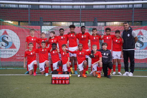 Eine Gruppe junger Männer in roten T-Shirts und schwarzen Shorts steht auf einem Fußballfeld neben einer Kiste roter Gegenstände, im Hintergrund Schilder mit "FC Red Black Eglaute - Champions League".