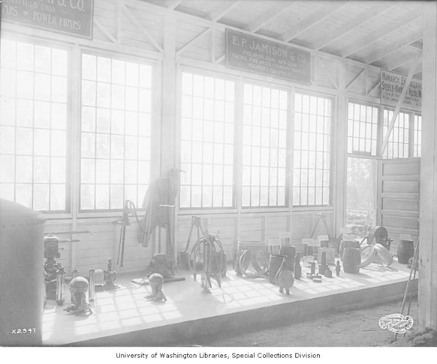 Schwarz-weiß-Foto eines großen Raums mit verschiedenen Werkzeugen und Maschinen auf dem Boden, Texttafeln an den Wänden und Glasfenstern, beschriftet mit "P.J. Jamison & Co. Power Pumps" unten.