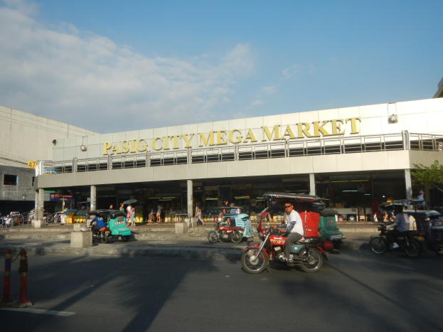 Eine belebte Straßenszene am Pasig City Mega Market mit Fahrzeugen und Fußgängern, ein Gebäude mit Text im Hintergrund unter einem bewölkten Himmel.