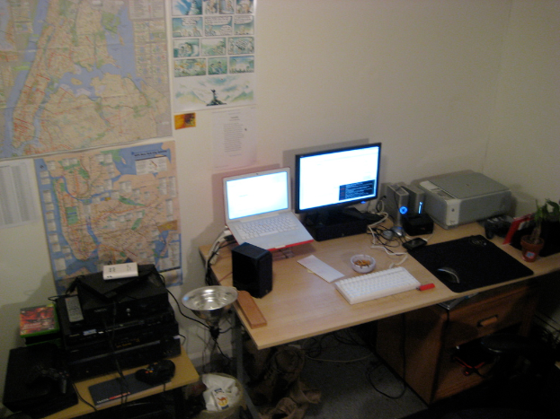Ein Raum mit einem Tisch, auf dem ein Laptop, Lautsprecher, Monitor, Tastatur, Ball, Maus, Scanner und Papiere stehen, sowie einem weiteren Tisch mit Elektronikgeräten im Hintergrund und Charts und Postern an der Wand.