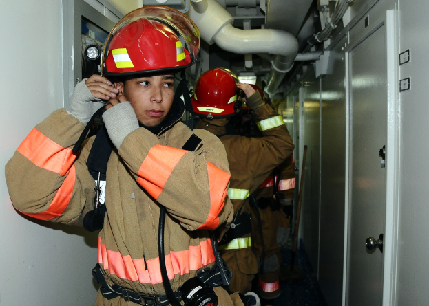 Feuerwehrleute in Uniform stehen in einem Raum mit einer Tür auf der rechten Seite und einer Wand auf der linken Seite während eines Trainings, mit Rohren im Hintergrund.