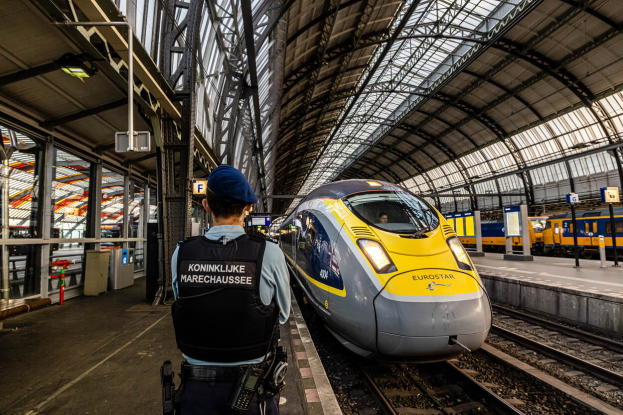 Ein Polizeibeamter in Uniform steht auf einem Bahnsteig neben einem Hochgeschwindigkeitszug, mit Menschen links, einem anderen Zug rechts und von der Decke beleuchteten Station.