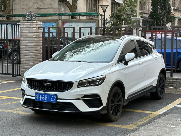 Ein weißer Volvo XC40 T5 AWD Inscription auf einem Parkplatz mit Gebäuden, Bäumen, Pfosten, Lampen und verschiedenen anderen Gegenständen im Hintergrund.