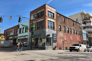 Ein großes Backsteingebäude an einer Straßenecke, mit Fahrzeugen, Fußgängern und Verkehrsampeln im Vordergrund und einem klaren Himmel im Hintergrund.