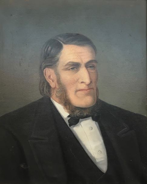 Portrait eines bärtigen Mannes in einem schwarzen Anzug und weißem Hemd mit ernstem Gesichtsausdruck, verschränkten Händen und aufrechter Haltung.