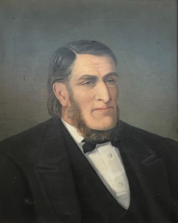 Portrait eines bärtigen Mannes in einem schwarzen Anzug und weißem Hemd mit ernstem Gesichtsausdruck, verschränkten Händen und aufrechter Haltung.