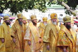 Eine Gruppe von Priestern in gelben Roben und Hüten, die eine Straße entlanggehen, umgeben von Menschen mit Schirmen, mit einem Gebäude und einem Baum im Hintergrund.