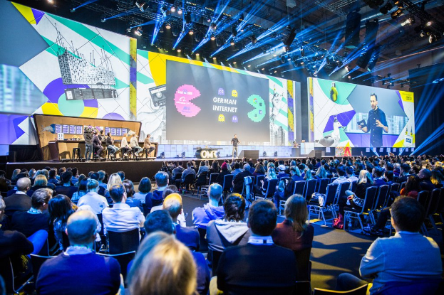 Eine große Menschenmenge sitzt auf Stühlen gegenüber einer Bühne bei der Deutschen Internetkonferenz 2019, mit einem Tisch voller Gegenstände links und Bildschirmen und Lichtern im Hintergrund.