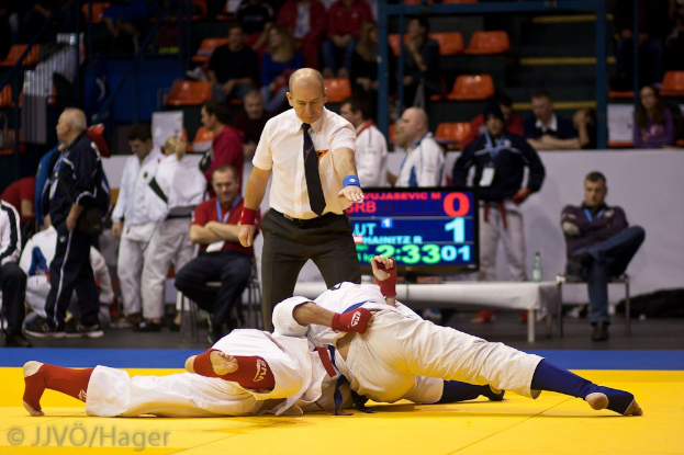 Zwei Judokämpfer im Kampf, mit einem Schiedsrichter in der Nähe, in einer Halle mit sitzenden und stehenden Zuschauern, Geländern, Treppen und einer Anzeigetafel im Hintergrund.