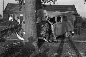 Ein Schwarz-Weiß-Bild eines verunglückten Autos am Straßenrand, mit einem Baum im Vordergrund, einigen Menschen in der Nähe und Häusern, Bäumen und einem Pfahl im Hintergrund.