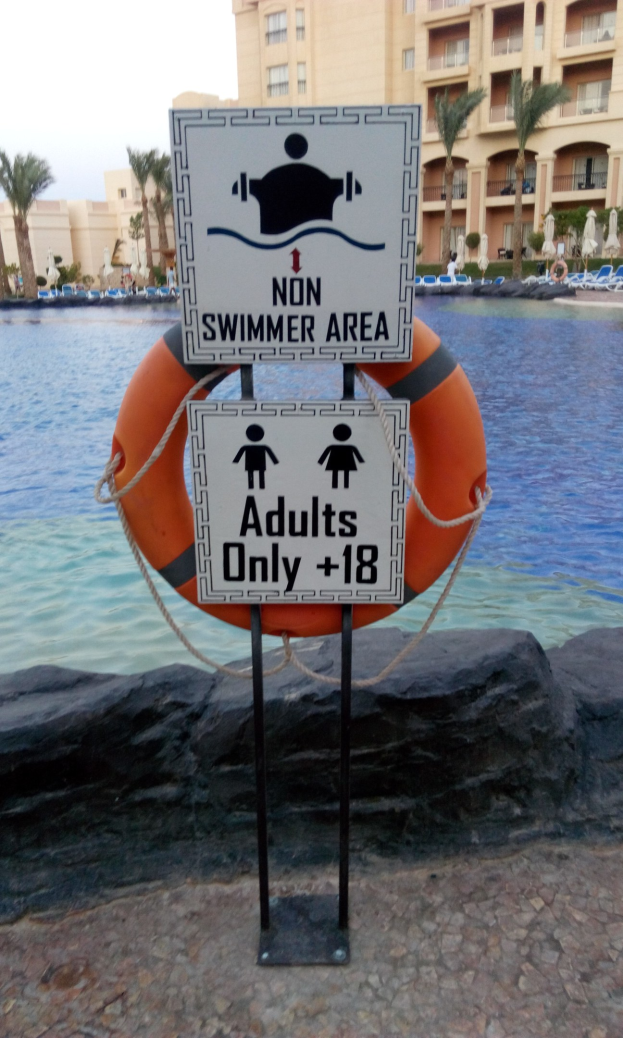 Ein Schwimmbad mit einem "Nur für Erwachsene - Nichtschwimmerbereich"-Schild im Vordergrund, umgeben von Bäumen und Gebäuden unter einem klaren blauen Himmel, mit einem Rettungsring in der Mitte des Pools.