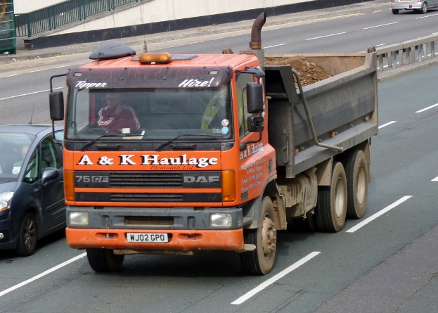 Ein Lastwagen mit einer Person darin ist in der Mitte, mit einem Auto mit einer Person links, einer Straße unten und einem Zaun, einem anderen Auto, Geländer und Wand im Hintergrund.
