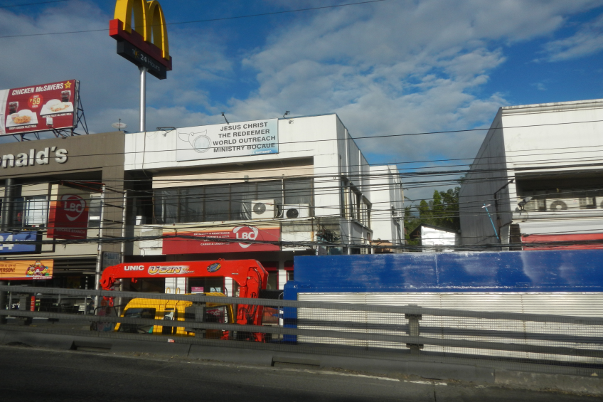 Ein McDonald's-Restaurant an einer Straßenecke, umgeben von Gebäuden, Werbetafeln, Strommasten, Fahrzeugen, Geländern, Bäumen und einem bewölkten Himmel.