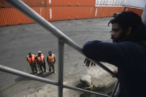 Ein Mann in legerer Kleidung steht auf einem Metallgeländer in der Nähe einer Gruppe von Menschen mit orangen Jacken und Kappen, mit Containern und einer Wand im Hintergrund am Hafen von Los Angeles.