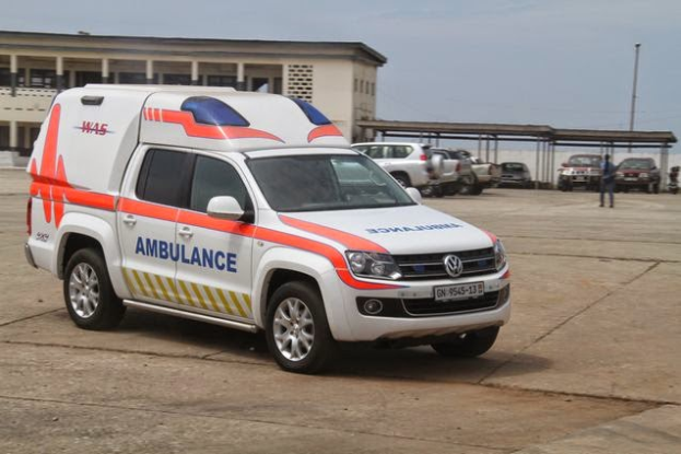 Ein Rettungswagen steht vor einem Gebäude mit einer Person in der Nähe, Masten säumen die Straße und ein Himmel ist im Hintergrund zu sehen, wahrscheinlich im Zusammenhang mit dem Nigerianischen Roten Kreuz, das medizinische Hilfe leistet.