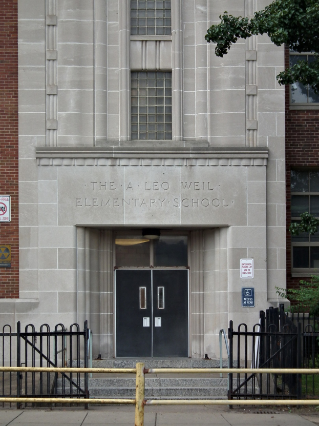 Außenansicht der Leo Weil Grundschule mit Fenstern, Türen, Schildern, einem Metallzaun, einem Weg, Gras, Pflanzen und einem Baum.