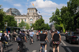 Eine Gruppe von Menschen, die auf Fahrrädern eine von Bäumen gesäumte Straße in Berlin, Deutschland, entlangfahren, mit Gebäuden auf beiden Seiten, einer Bushaltestelle auf der rechten Seite und einer Flagge, die auf einem der Gebäude weht, unter einem bewölkten Himmel.